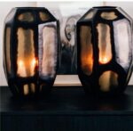 Twee decoratieve vazen met geometrische patronen staan op een donker oppervlak. Elke vaas heeft een warm, gloeiend licht binnenin. Een zwart-witportret van een persoon met een open mond is gedeeltelijk zichtbaar op de achtergrond.