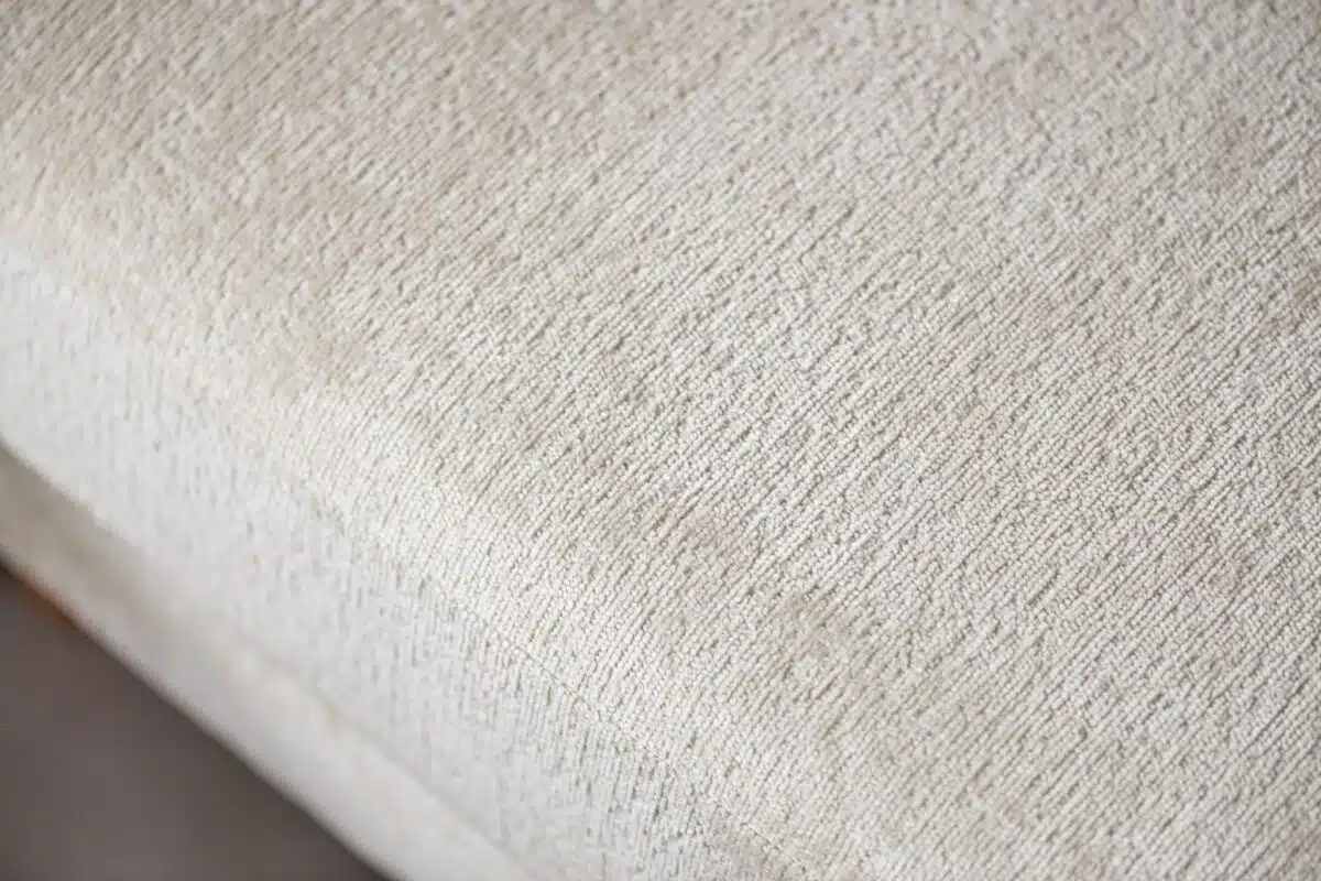 Close-up van een lichtbeige textuurstof met een subtiel geweven patroon. Het materiaal lijkt glad met een lichte glans, waardoor de ingewikkelde details van het weefsel worden benadrukt.