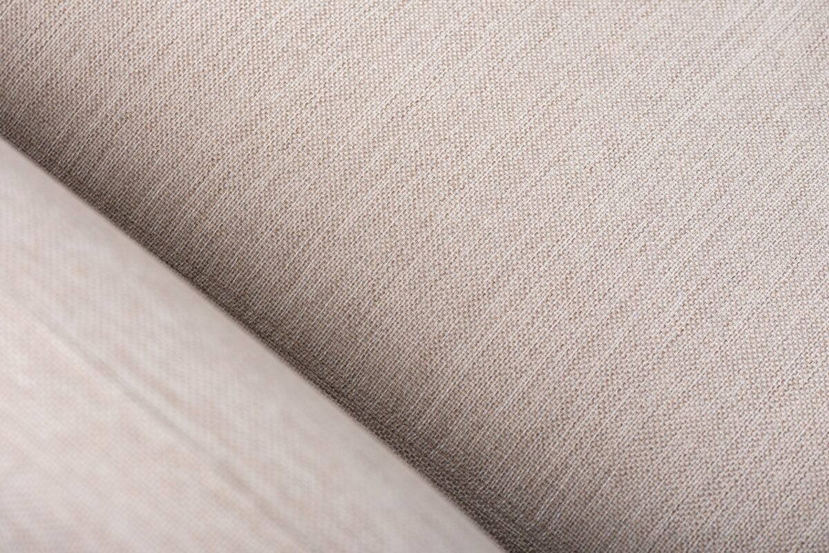Close-up van beige textuurstof met een diagonale naad. De stof heeft een geweven uiterlijk en de naad creëert een subtiele verdeling in de afbeelding, waardoor de details en textuur van het materiaal worden benadrukt.
