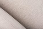 Close-up van beige textuurstof met een diagonale naad. De stof heeft een geweven uiterlijk en de naad creëert een subtiele verdeling in de afbeelding, waardoor de details en textuur van het materiaal worden benadrukt.