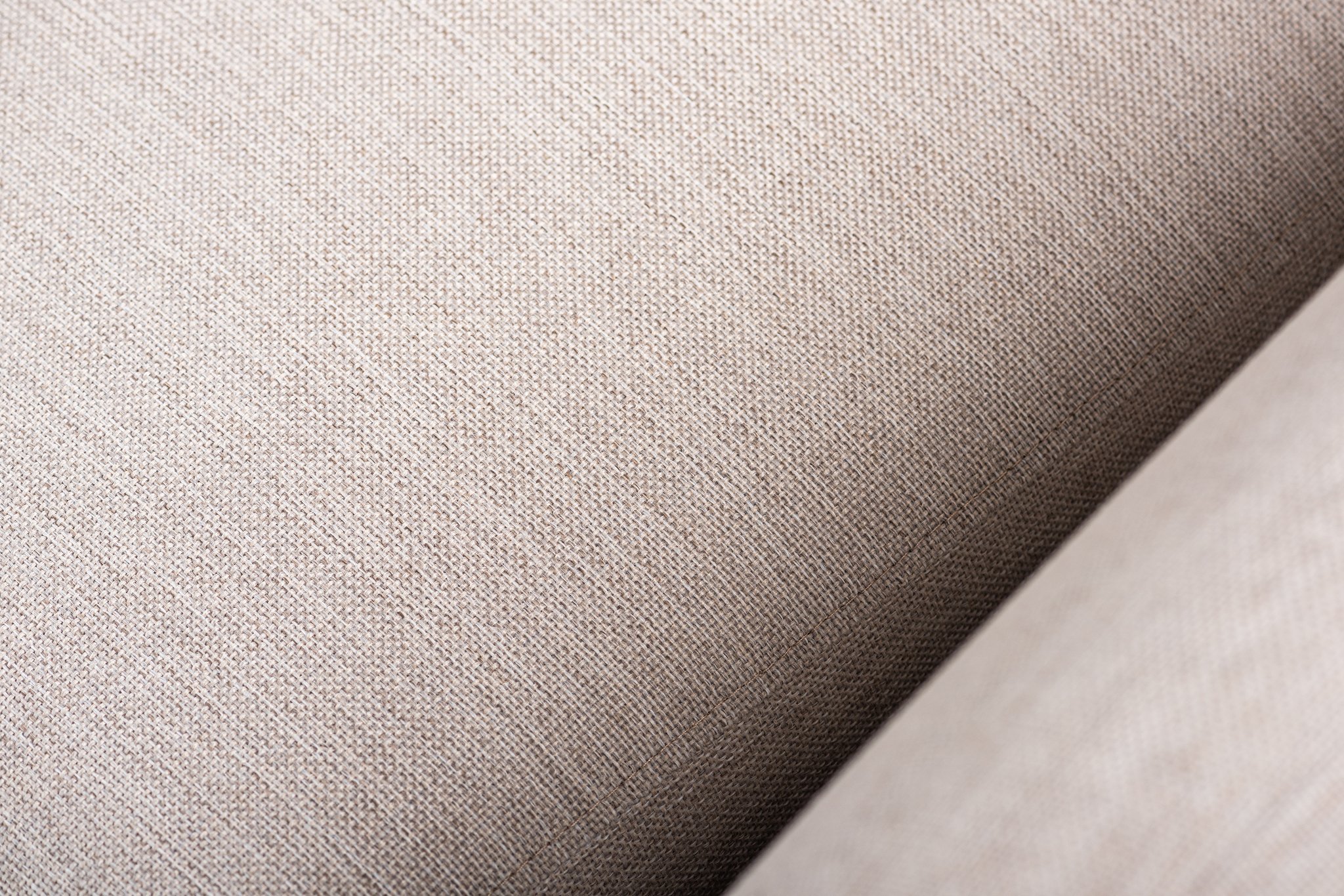 Close-up van een beige stoffentextuur met een diagonale oriëntatie. Het oppervlak vertoont een fijn weefpatroon, wat het een enigszins ruw uiterlijk geeft. De belichting benadrukt de subtiele variaties in toon en textuur van de stof.