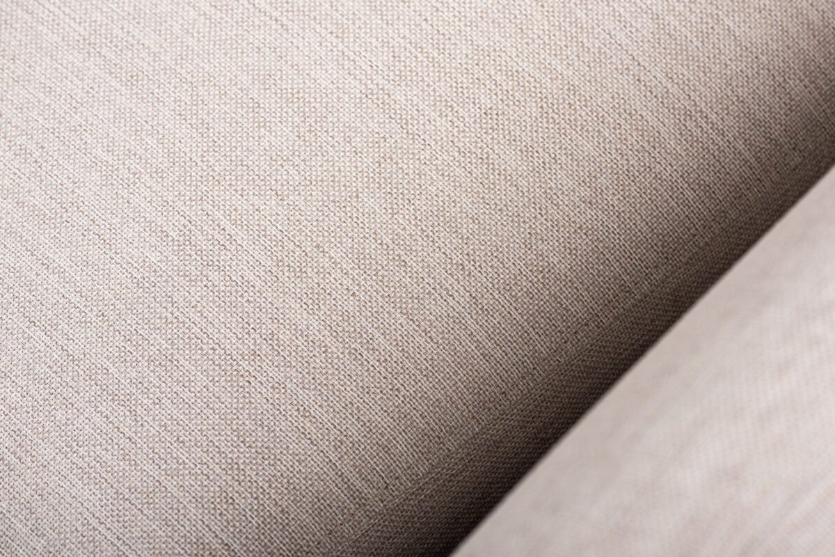 Close-up van beige stof met een fijn geweven structuur. Het materiaal lijkt glad, met subtiele variaties in schaduw, wat een zacht en comfortabel gevoel suggereert. De afbeelding richt zich op de details van de vezels van de stof.
