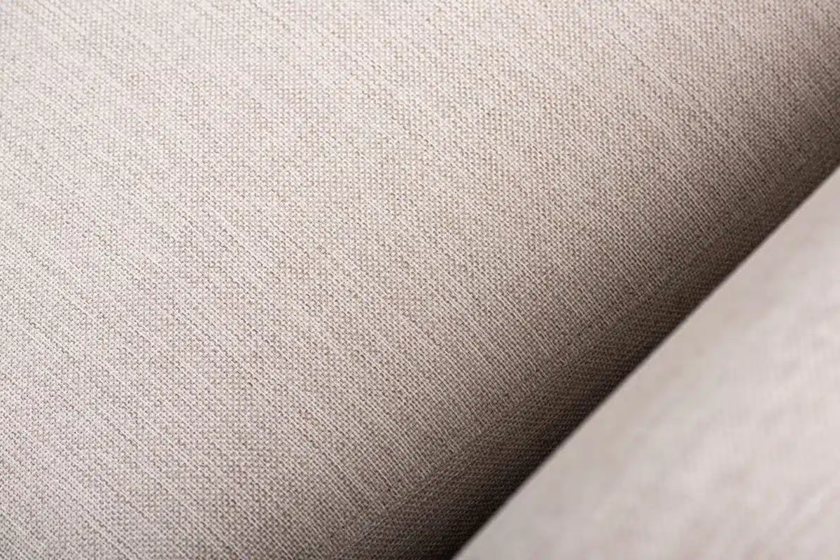 Close-up van beige stof met een fijn geweven structuur. Het materiaal lijkt glad, met subtiele variaties in schaduw, wat een zacht en comfortabel gevoel suggereert. De afbeelding richt zich op de details van de vezels van de stof.