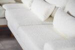 Close-up van een moderne, witte, getextureerde sectionele bank met pluche kussens. De stof heeft een zachte, donzige uitstraling. De bank staat op een gladde, lichtgekleurde vloer en de afbeelding richt zich op de textuur en het ontwerp van het zitgedeelte.