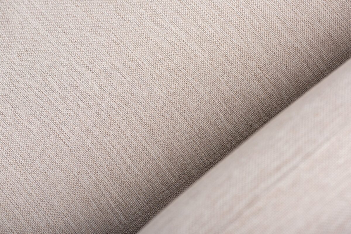Close-up van een lichtbeige stof met een subtiele, geweven textuur. Het oppervlak toont fijne draden die horizontaal en verticaal lopen, waardoor een eenvoudig maar elegant patroon ontstaat.
