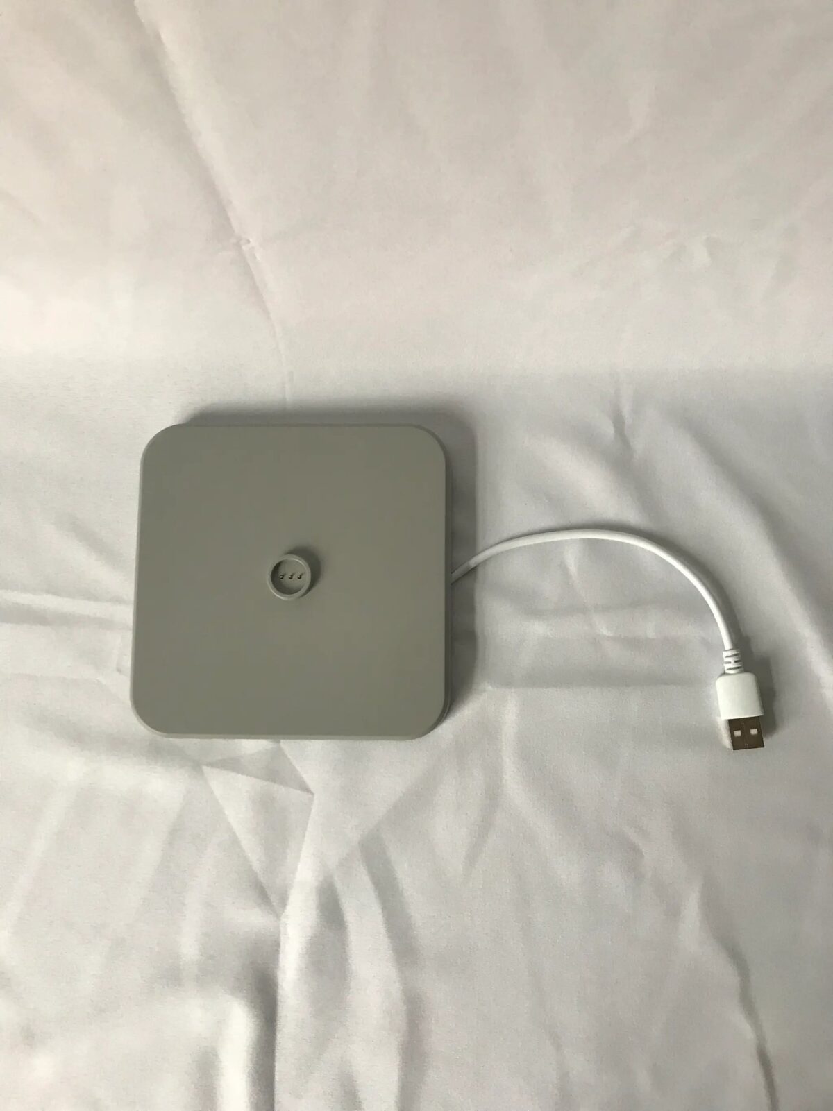 Een vierkante grijze oplaadpad met een gecentreerde knop ligt op een wit stoffen oppervlak. Een korte witte USB-kabel is bevestigd aan de zijkant van de pad.