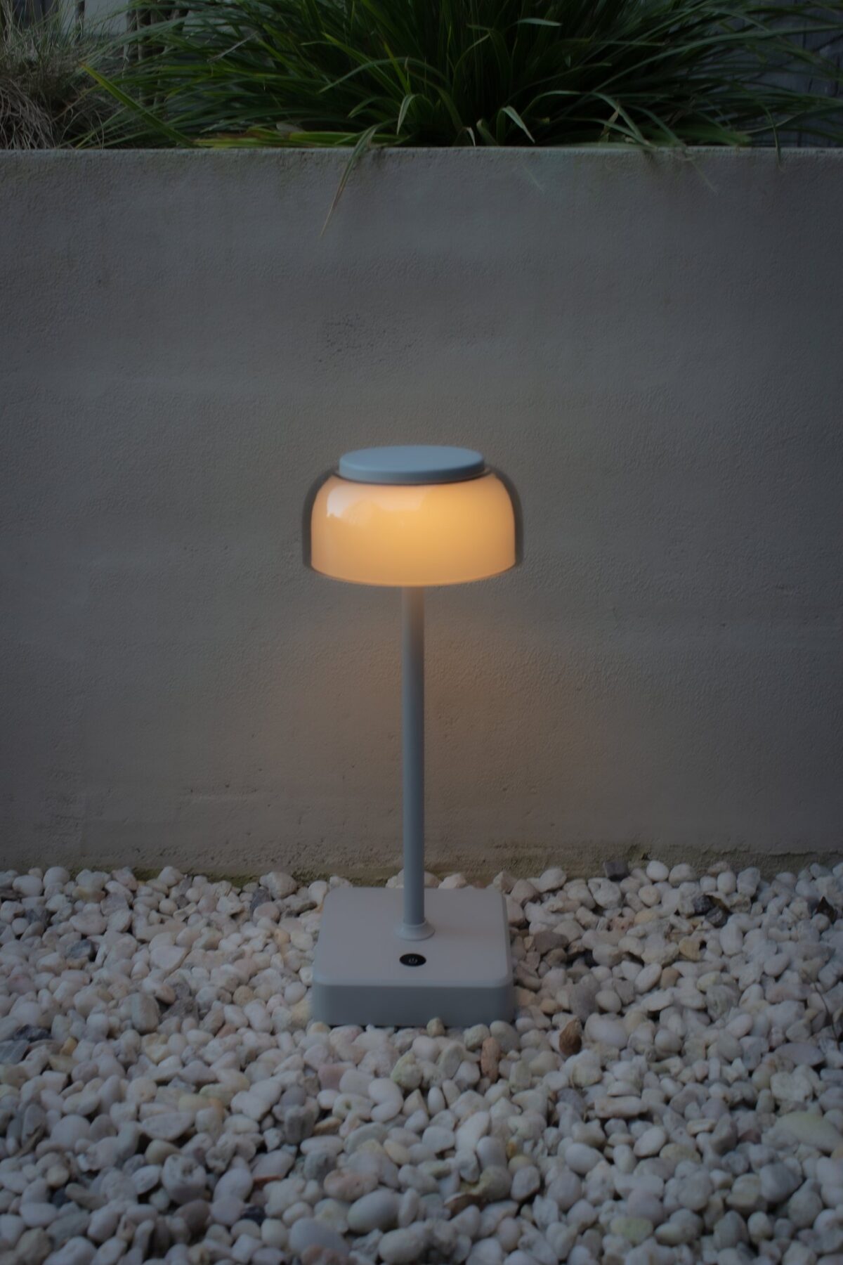 Een moderne, minimalistische buitenlamp met een ronde bovenkant straalt een warme gloed uit. Hij staat op een vierkante basis omringd door witte kiezels. Op de achtergrond is er een minimalistische betonnen muur en groene planten.