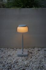 Een moderne, minimalistische buitenlamp met een ronde bovenkant straalt een warme gloed uit. Hij staat op een vierkante basis omringd door witte kiezels. Op de achtergrond is er een minimalistische betonnen muur en groene planten.