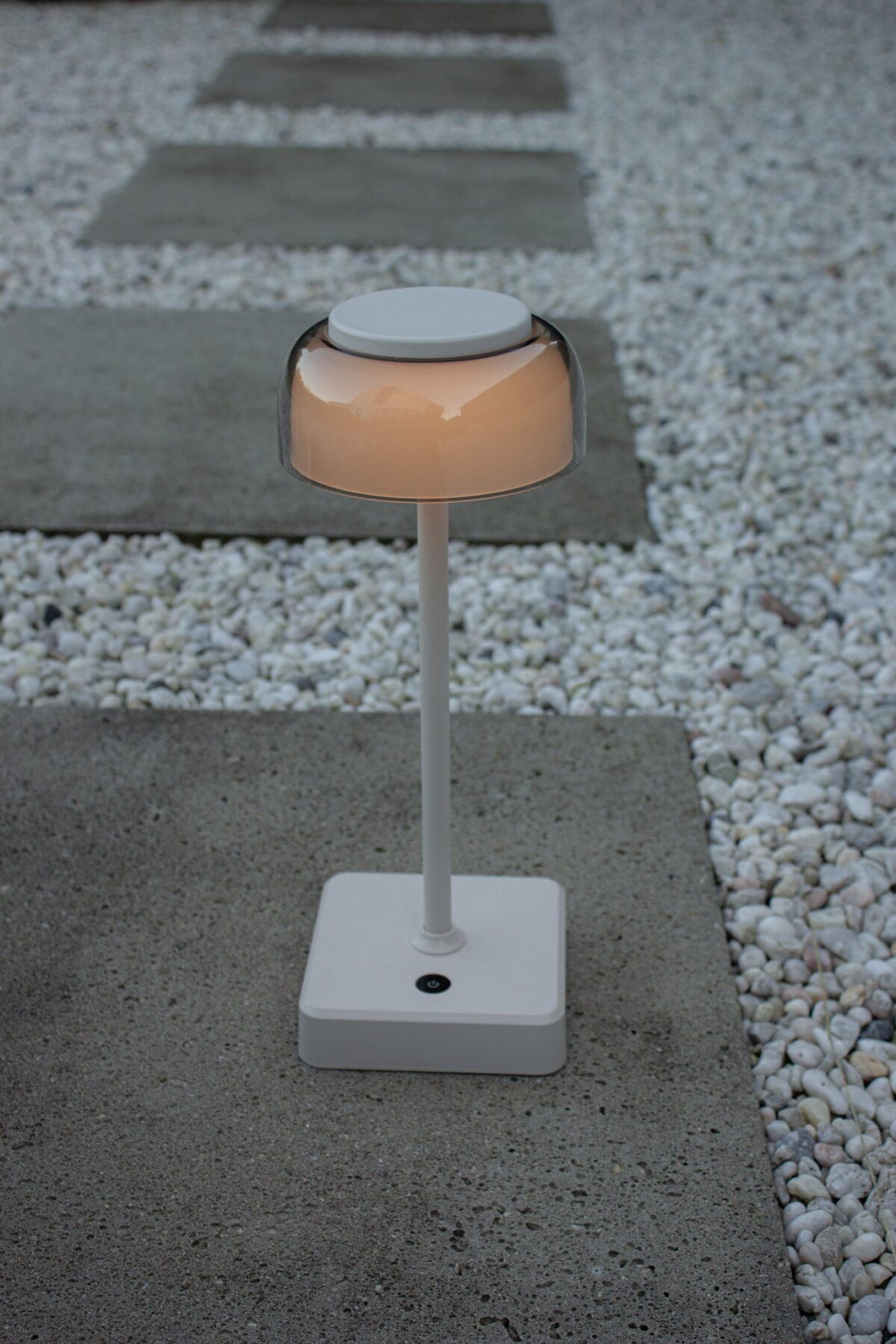 Een moderne, cilindervormige buitenlamp met een transparant bovendeel en een witte voet, geplaatst op een betonnen plaat, omringd door witte kiezels en rechthoekige betonnen stapstenen.