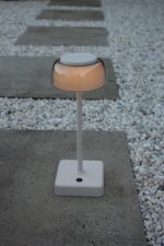 Een moderne, cilindervormige buitenlamp met een transparant bovendeel en een witte voet, geplaatst op een betonnen plaat, omringd door witte kiezels en rechthoekige betonnen stapstenen.
