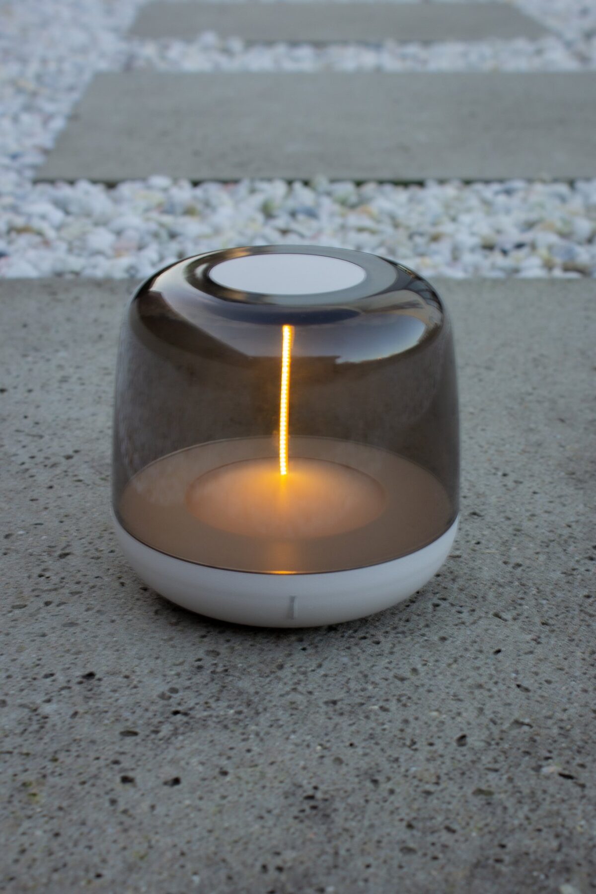 Een moderne kaarsenwarmer met een doorschijnende, koepelvormige kap en een witte basis staat op een betonnen oppervlak. Het apparaat straalt een warm, gloeiend licht uit. Op de achtergrond is een pad met witte stenen te zien.