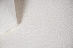 Close-up van een getextureerde off-white stof met een hoek die lichtjes is omgevouwen. De stof lijkt zacht, met een subtiel patroon dat het een pluizige uitstraling geeft. De belichting benadrukt de textuur, wat diepte toevoegt aan de afbeelding.