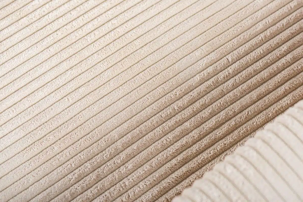 Close-up van een beige corduroystof met zijn getextureerde, geribbelde patroon. Het materiaal lijkt zacht, met parallelle lijnen die diagonaal over de afbeelding lopen, wat de pluche en tastbare kwaliteit benadrukt.