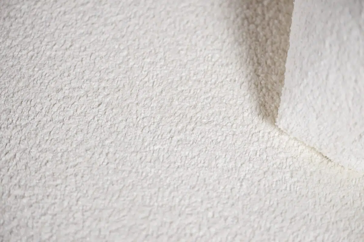 Close-up van een getextureerd wit oppervlak met een hoek die iets is opgetild, waardoor een gladde onderkant zichtbaar wordt. De belichting benadrukt de ruwe textuur, waardoor subtiele schaduwen en een gevoel van diepte ontstaan.