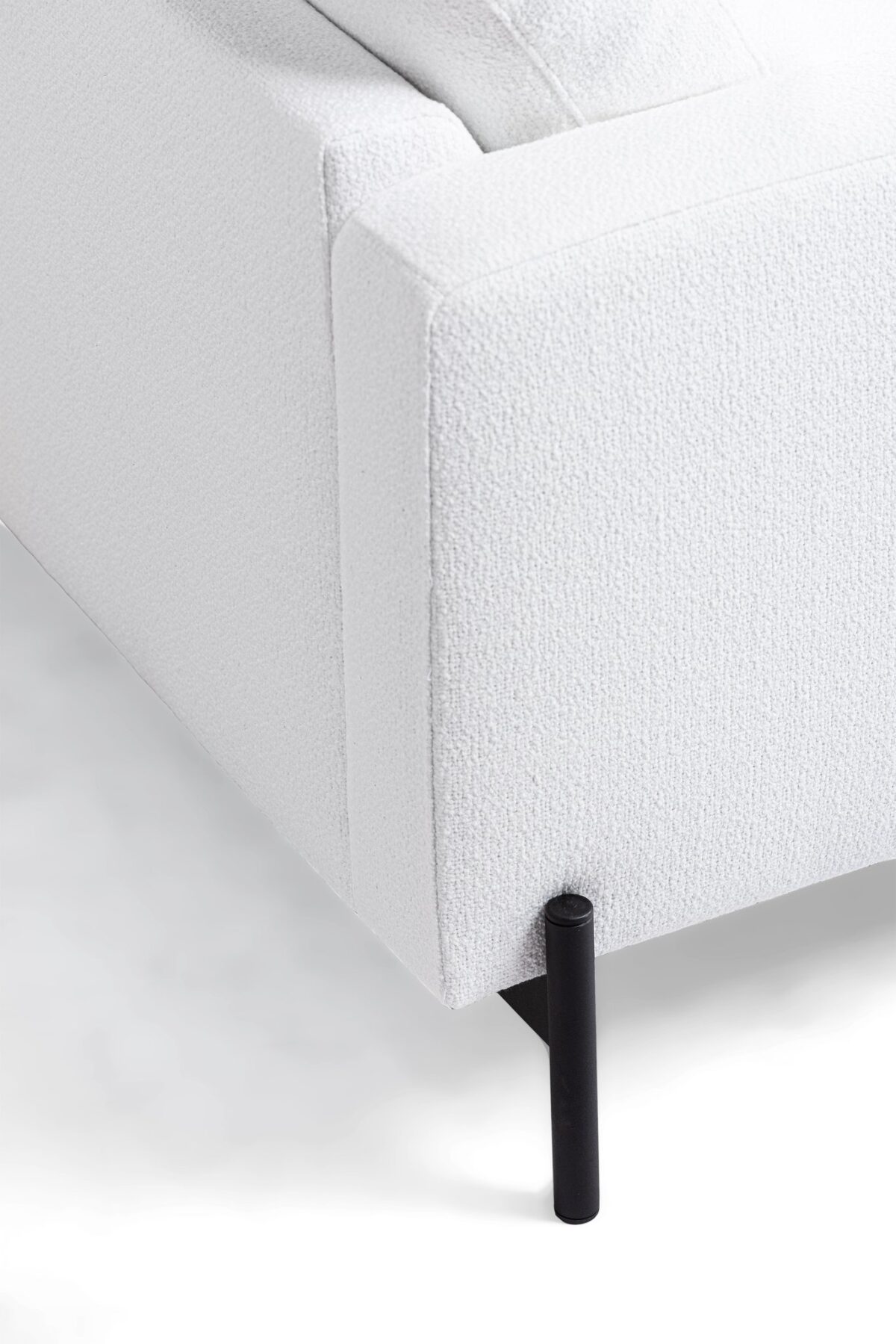 Close-up van de hoek van een witte getextureerde bank met minimalistische zwarte metalen poten. De stof oogt zacht en heeft een licht ruwe textuur. De strakke lijnen geven het een moderne, elegante look tegen een neutrale achtergrond.