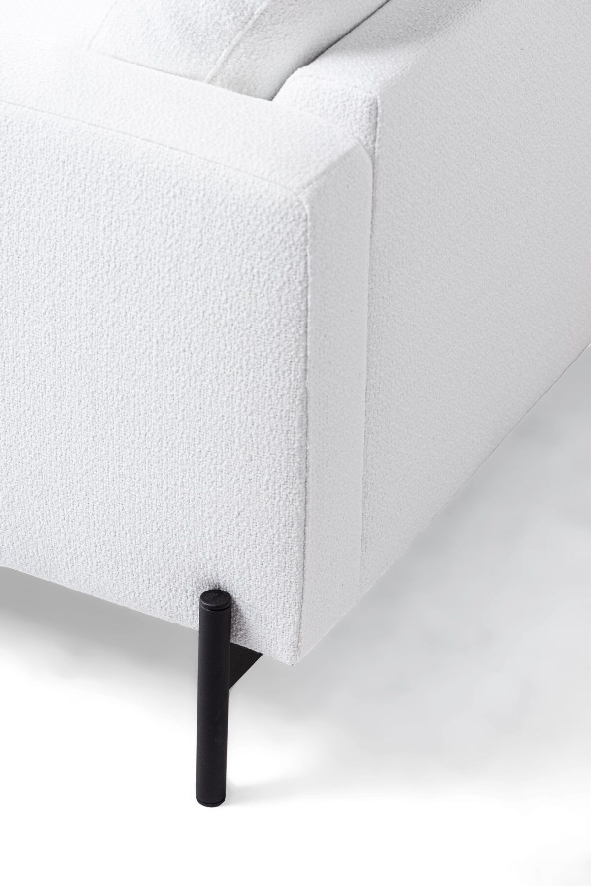 Close-up van een moderne hoekbank in witte stof. De armleuning en de rugleuning zijn zichtbaar, met een gestructureerd oppervlak. De bank staat op strakke, cilindrische zwarte poten, wat het minimalistische ontwerp benadrukt. Het oppervlak waarop de bank staat, lijkt wit.