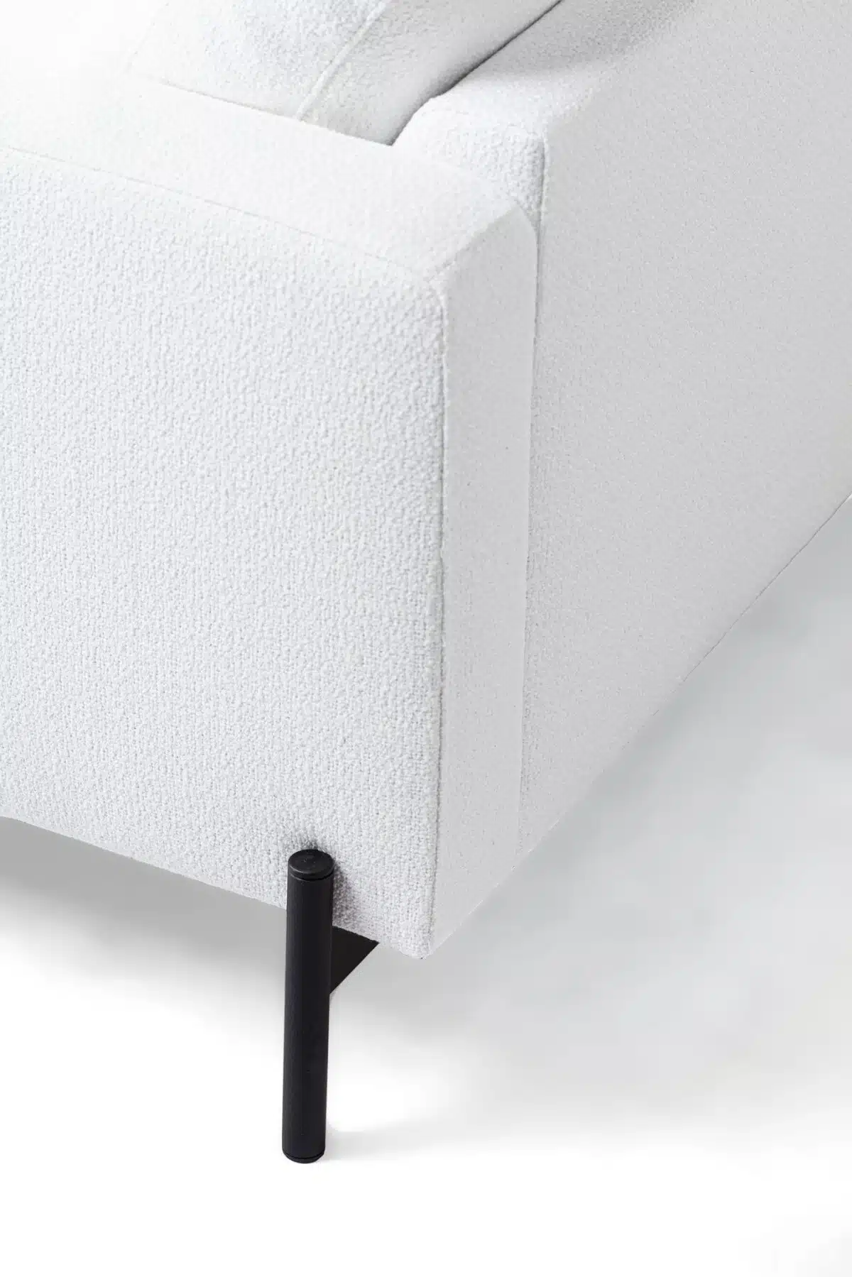 Close-up van een moderne hoekbank in witte stof. De armleuning en de rugleuning zijn zichtbaar, met een gestructureerd oppervlak. De bank staat op strakke, cilindrische zwarte poten, wat het minimalistische ontwerp benadrukt. Het oppervlak waarop de bank staat, lijkt wit.