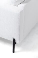 Close-up van een moderne hoekbank in witte stof. De armleuning en de rugleuning zijn zichtbaar, met een gestructureerd oppervlak. De bank staat op strakke, cilindrische zwarte poten, wat het minimalistische ontwerp benadrukt. Het oppervlak waarop de bank staat, lijkt wit.