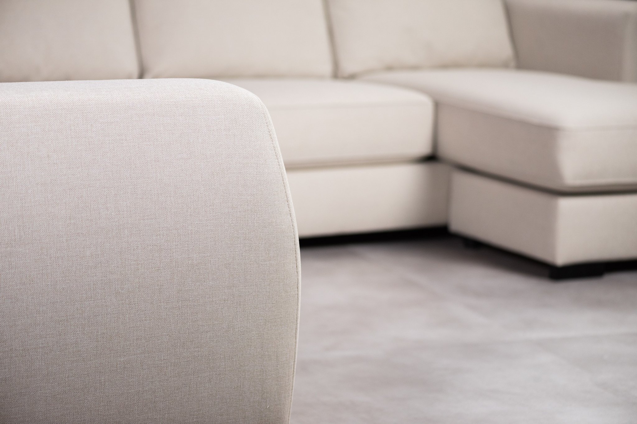 Close-up van een beige hoekbank met stoffen bekleding. De focus ligt op de armleuning op de voorgrond, terwijl de rest van de L-vormige bank enigszins wazig is op de achtergrond, afgezet tegen een bijpassend beige tapijt.