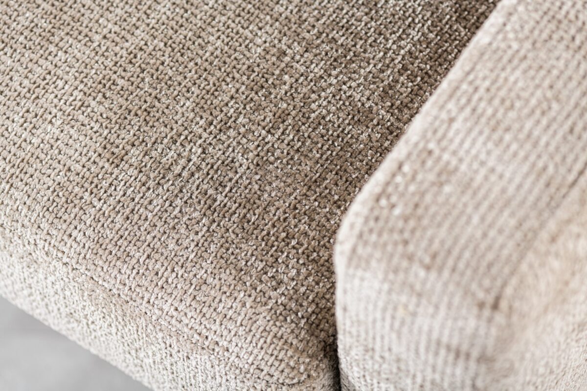 Close-up van een getextureerde stoffen bekleding op een beige bank. De afbeelding legt het weefpatroon vast en de hoek waar de armleuning het zitkussen raakt.