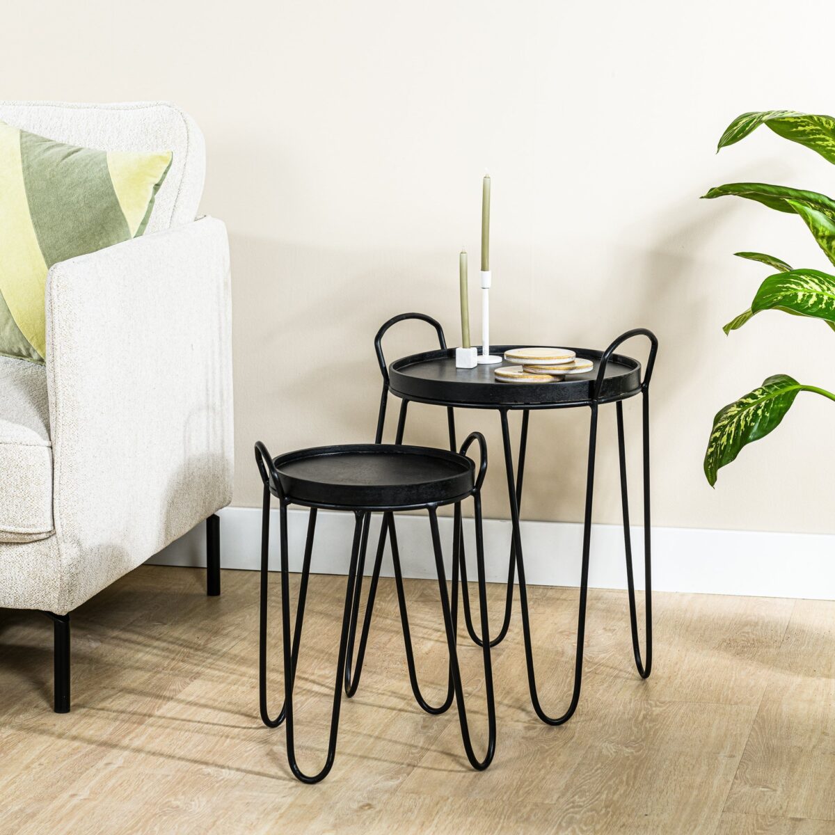 Twee Salontafels, set van 2, A340 zwart met haarspeldpoten staan op een lichthouten vloer bij een beige bank met groene kussens. Op één tafeltje staan twee groene kaarsen en onderzetters, met een bladplant in de buurt.