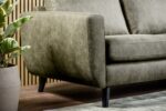 Een close-up van de 3 zits bank CL L+R in S156 groen Savannah stof, met houten poten, op een vloerkleed naast een potplant en houten latten wand.