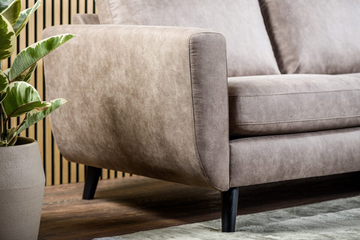 Een close-up van de 3 zits bank CL L+R in Savannah S520 taupe stof, met zwarte poten, naast een groene potplant op een houten vloer, met een deel van een vloerkleed met textuur en verticale houten latten op de achtergrond.