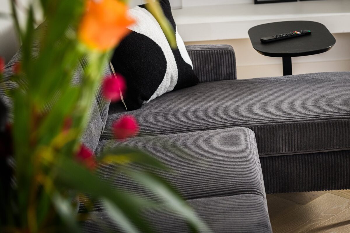 Een antraciet 3-zitsbank (CL L+R, stof Rib230) staat naast een klein zwart bijzettafeltje met een tv-afstandsbediening; een zwart-wit kussen rust op de bank en op de voorgrond zijn vage bloemen te zien.