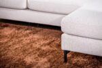 Close-up van een moderne lichtbeige sectionele bank op een pluche bruin tapijt. De bank heeft een strak, minimalistisch design met metalen poten, wat contrasteert met de zachte textuur van het tapijt.