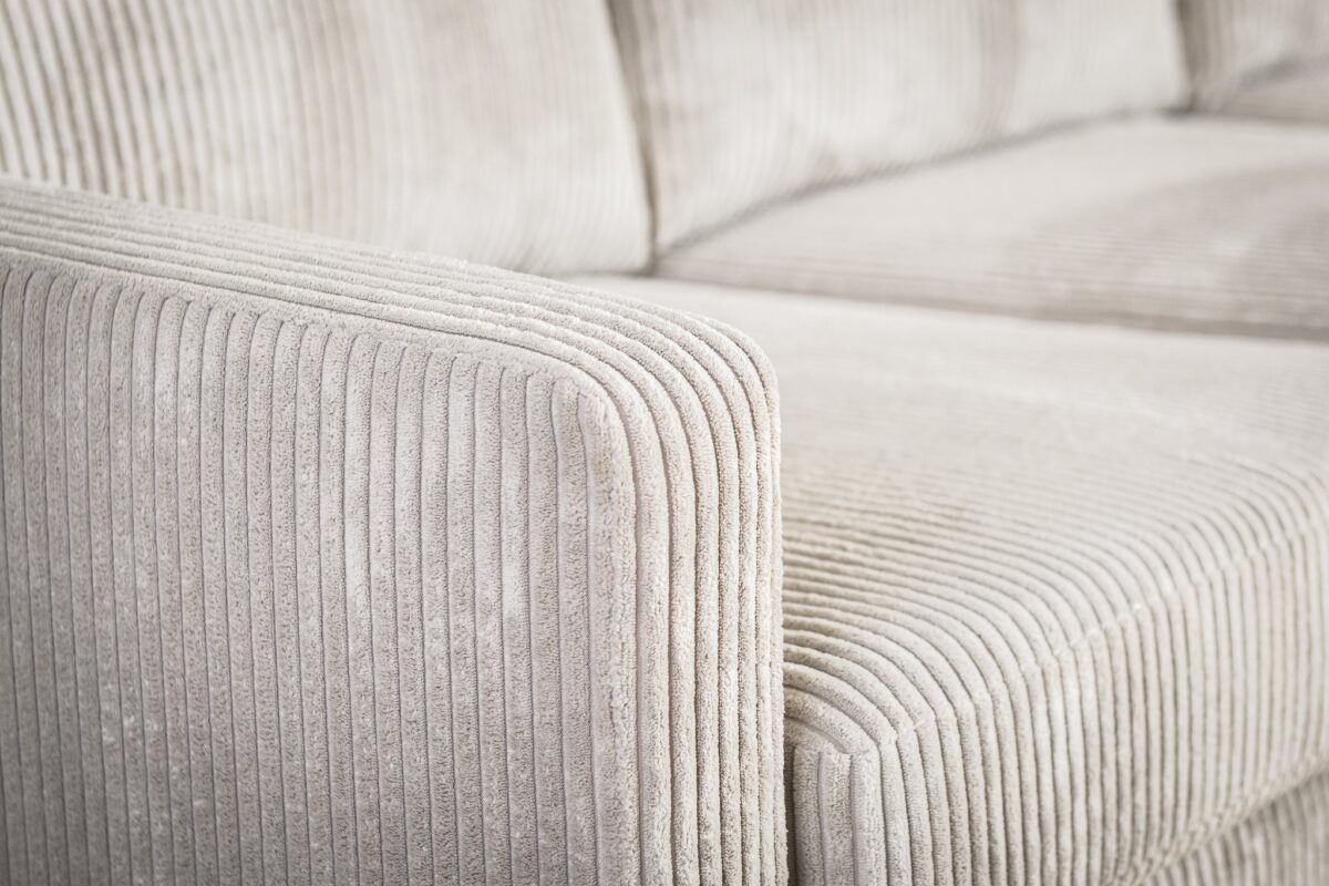 Close-up van een beige corduroy bank, met de structuurstof en zachte, geribbelde lijnen. De afbeelding richt zich op de armleuning en een deel van het zitgedeelte, wat de pluche en uitnodigende uitstraling van de bank benadrukt.