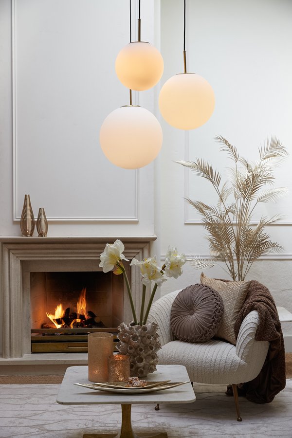 Een gezellige woonkamer met een verlichte open haard, een pluchen fauteuil en een tafeltje. Daarboven hangen drie bolvormige hanglampen. De kamer is versierd met een vaas met witte bloemen en pampasgras, wat een warme en uitnodigende sfeer creëert.
