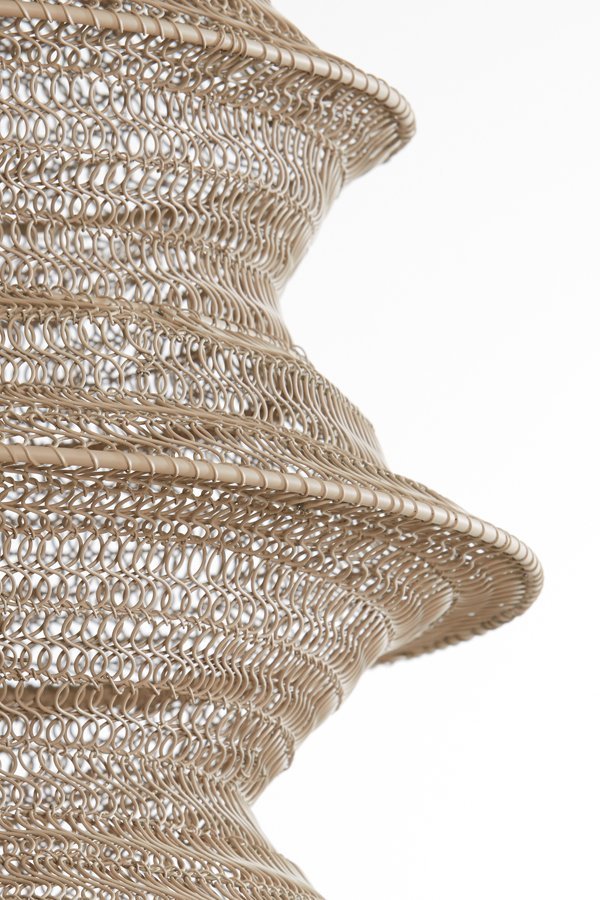 Close-up van een ingewikkelde, spiraalvormige geweven structuur van dun, lichtbruin materiaal. Het ontwerp bevat herhalende lussen en rondingen, waardoor een gestructureerde en luchtige uitstraling ontstaat tegen een effen witte achtergrond.