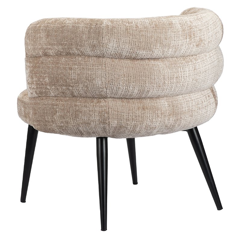 Een moderne beige gestoffeerde stoel met een ronde rugleuning en gelaagde kussens. De stoel heeft vier slanke zwarte taps toelopende poten. Hij is in een hoek geplaatst, waardoor de structuurstof en het eigentijdse ontwerp goed tot hun recht komen.