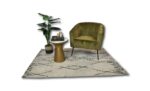 Een groene fluwelen fauteuil staat op een crèmekleurig geometrisch kleed. Een gouden bijzettafeltje bevat een kleine plant, kaars en boeken. Een hoge potplant staat naast de stoel, wat een gezellige, moderne opstelling creëert.
