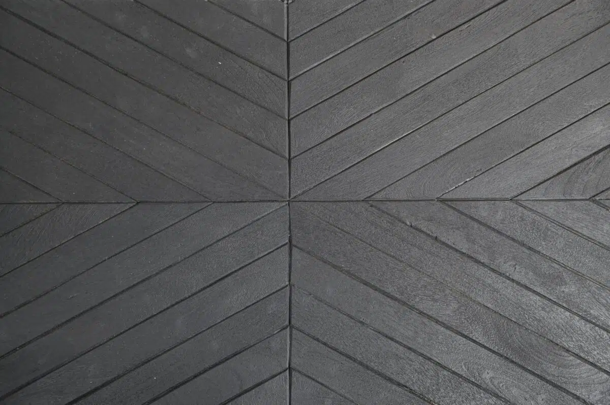 Donkere houten vloerplanken gerangschikt in een visgraatpatroon, convergerend in het midden in een symmetrisch ontwerp. Het hout heeft een gestructureerd oppervlak met zichtbare nerf en lichte variaties in kleur, wat een dynamisch visueel effect creëert.