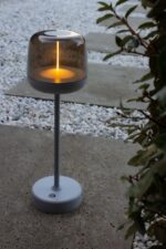 Een moderne tafellamp met een cilindrische voet en een rookglazen kap staat op een grindpad. De lamp straalt een warme, verticale gloed uit. In de buurt is wat groen gebladerte zichtbaar aan de rechterkant van de afbeelding.