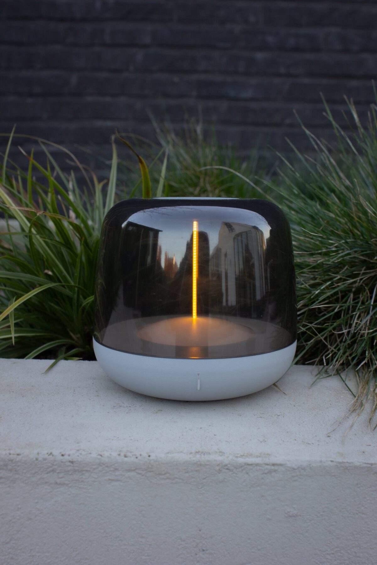 Een moderne, transparante lamp met een cilindrisch ontwerp en een gloeiend verticaal licht staat op een betonnen oppervlak. Daarachter is groen gras en een donkere bakstenen muur, die stedelijke architectuur in het oppervlak van de lamp weerspiegelen.
