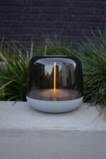 Een moderne, transparante lamp met een cilindrisch ontwerp en een gloeiend verticaal licht staat op een betonnen oppervlak. Daarachter is groen gras en een donkere bakstenen muur, die stedelijke architectuur in het oppervlak van de lamp weerspiegelen.