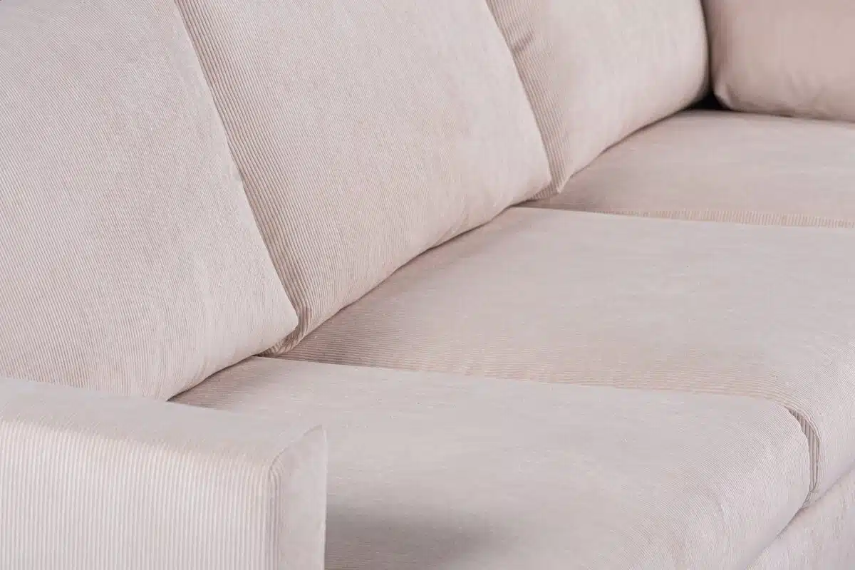 Close-up van een beige stoffen bank met drie zitkussens en twee armleuningen. Het materiaal heeft een subtiele gestreepte textuur en de kussens lijken zacht en comfortabel. De verlichting benadrukt de textuur en neutrale kleur van de bekleding.