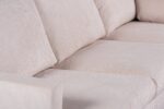 Close-up van een beige stoffen bank met drie zitkussens en twee armleuningen. Het materiaal heeft een subtiele gestreepte textuur en de kussens lijken zacht en comfortabel. De verlichting benadrukt de textuur en neutrale kleur van de bekleding.