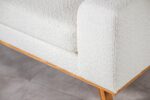 Close-up van een witte structuurbank met zachte stof. De bank heeft een hardhouten basis en schuine houten poten, geplaatst op een grijze vloer. De bekleding heeft een bouclé-uitstraling, wat een gezellige en moderne touch toevoegt.