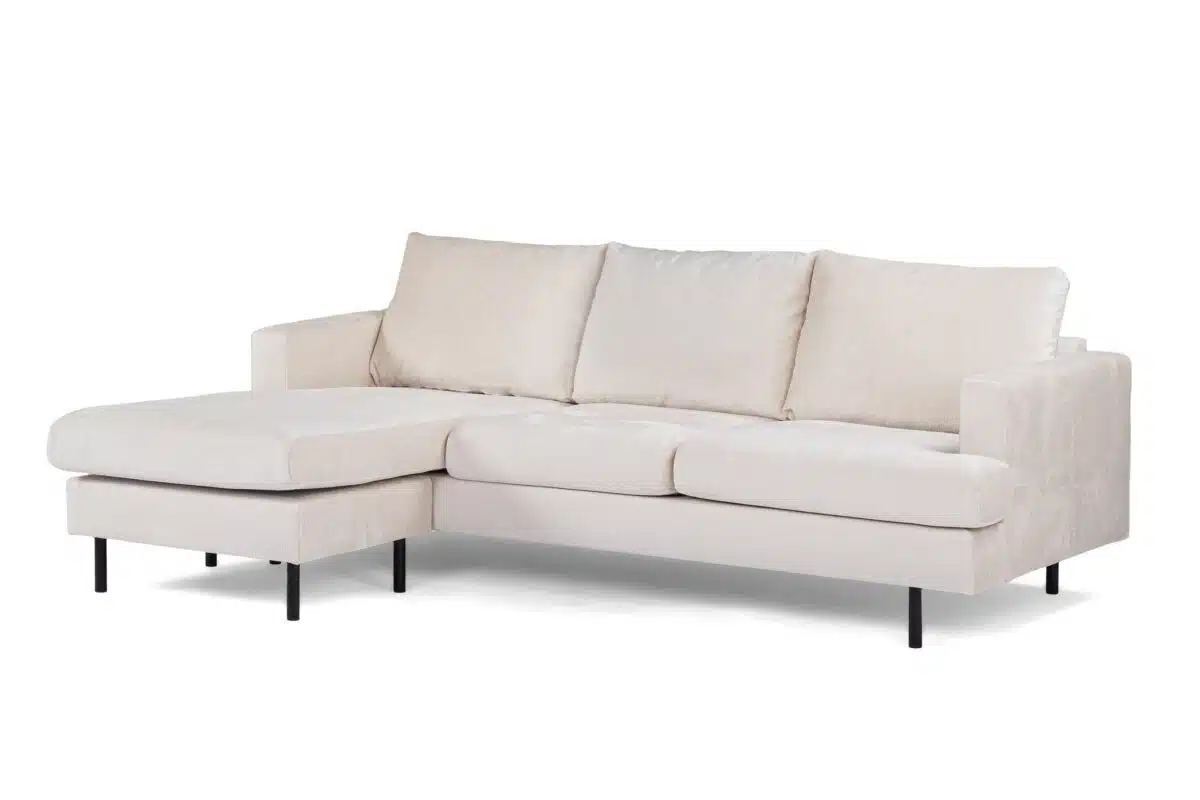 Crèmekleurige sectionele bank met drie kussens en een chaise longue aan de linkerkant. De bank heeft een minimalistisch ontwerp, wordt ondersteund door dunne zwarte poten en is bekleed met een zachte stof. Geplaatst tegen een effen witte achtergrond.