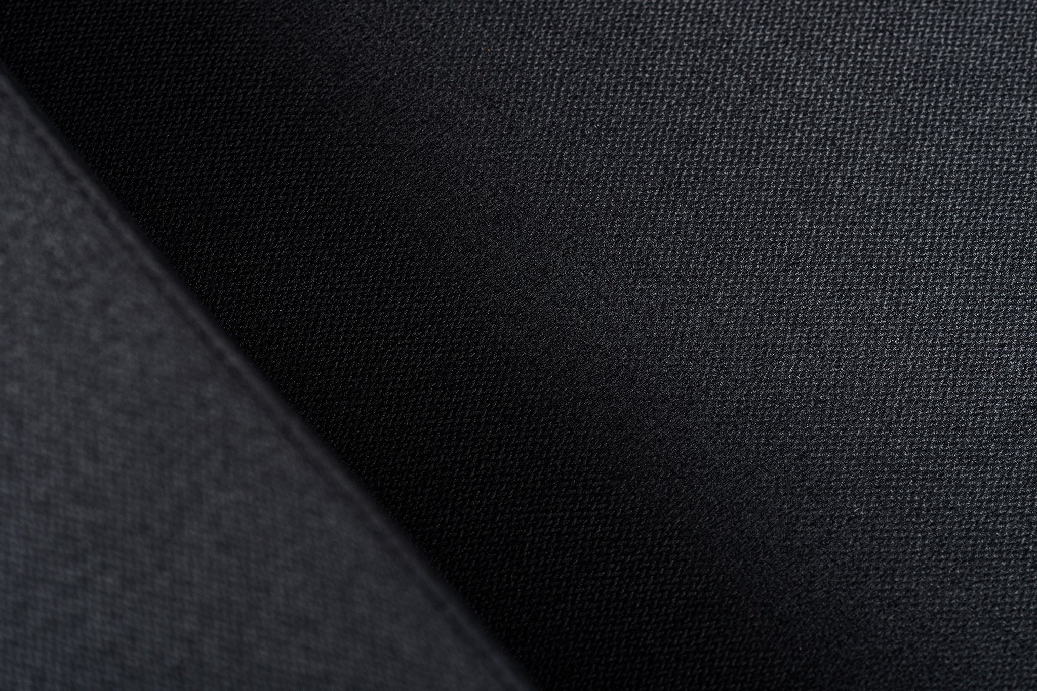 Close-up van een donkere textuurstof met een diagonale schaduw, die het subtiele weefpatroon en het gladde oppervlak benadrukt. Het licht creëert een zacht gradiënteffect over het materiaal.