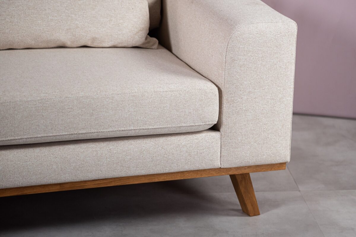 Een close-up van een beige gestoffeerde hoekbank met houten poten op een grijze tegelvloer. De rug- en zitkussens zijn pluche en licht getextureerd. De muur erachter is zacht mauvekleurig.