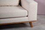 Een close-up van een beige gestoffeerde hoekbank met houten poten op een grijze tegelvloer. De rug- en zitkussens zijn pluche en licht getextureerd. De muur erachter is zacht mauvekleurig.