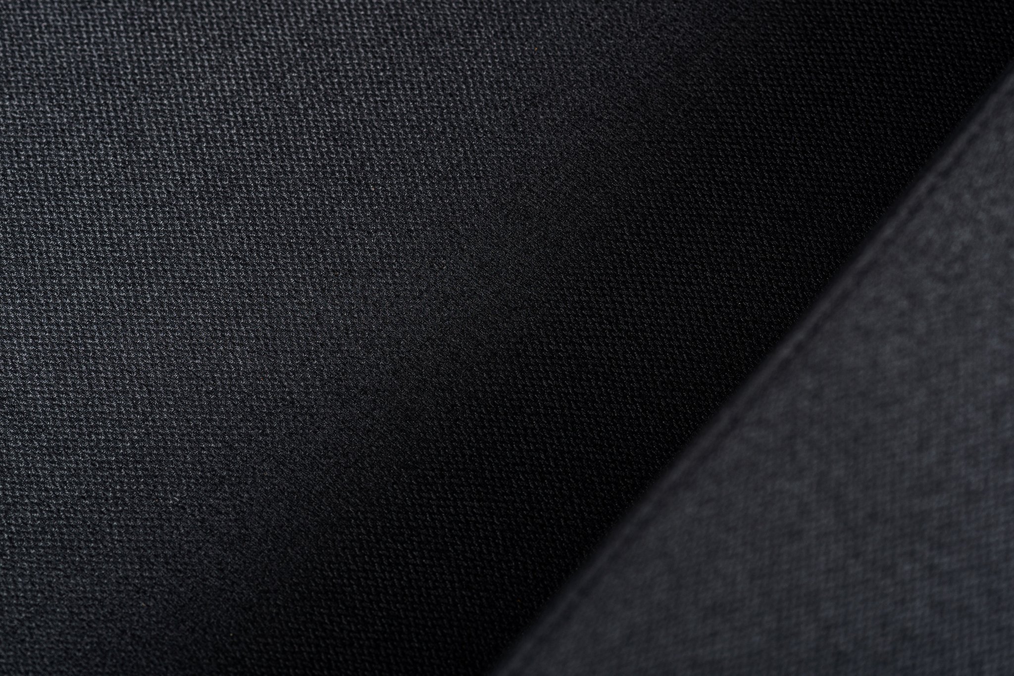 Close-up van een getextureerde zwarte stof met subtiele belichting. Het oppervlak toont een fijn weefpatroon, wat een gevoel van diepte en detail creëert. Schaduwen en highlights gaan subtiel over, wat de textuur en het uiterlijk van de stof versterkt.