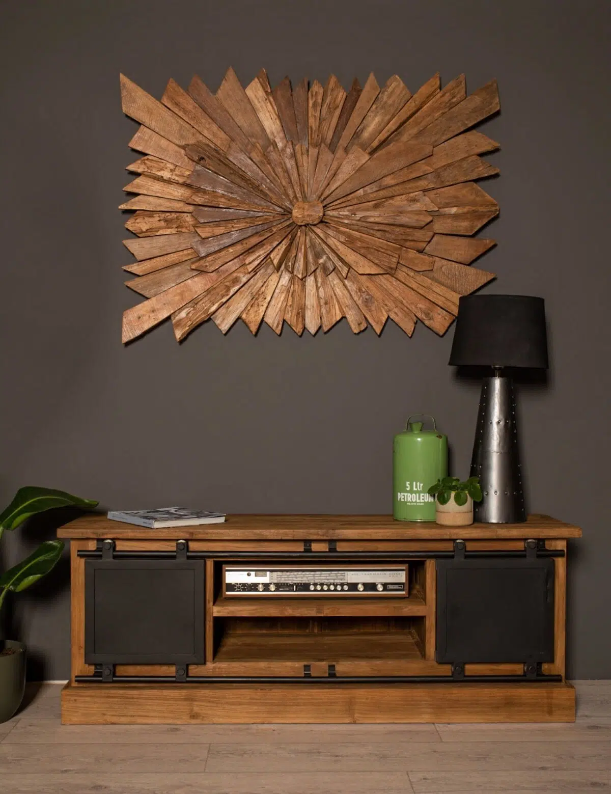 Houten dressoir met zwarte schuifpanelen, met een stereo. Een grote, stervormige houten wanddecoratie hangt erboven. Een hoge lamp en een groene gieter staan op het dressoir. Een plant met brede bladeren staat op de vloer. De muur is donkergrijs geverfd.
