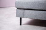 Close-up van een grijs gestoffeerde hoekbank met strakke zwarte metalen poten. De bank staat op een lichtgekleurde tegelvloer, met een minimalistische uitstraling en strakke lijnen.