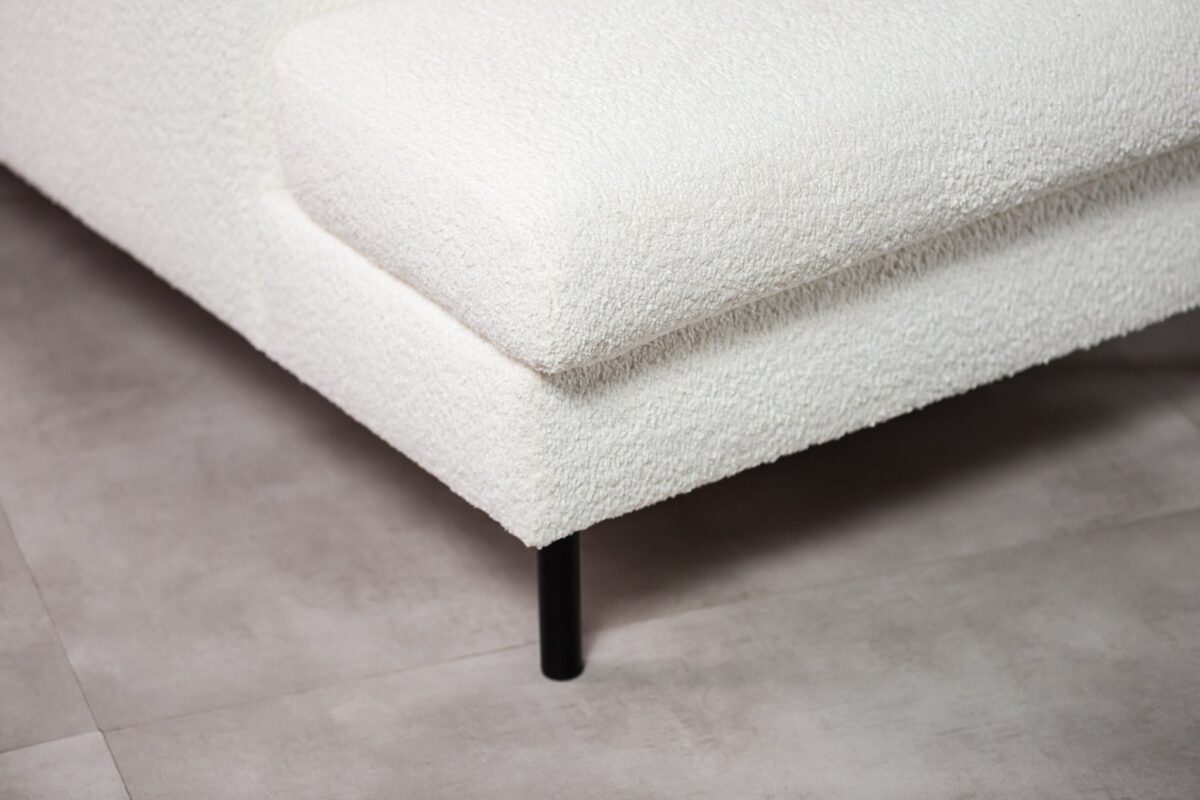 Close-up van een moderne witte getextureerde hoekbank met een zwarte metalen poot op een lichtgrijze betegelde vloer. De bekleding lijkt zacht en gestructureerd, in contrast met het strakke, minimalistische ontwerp van de poot.
