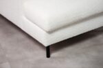 Close-up van een moderne witte getextureerde hoekbank met een zwarte metalen poot op een lichtgrijze betegelde vloer. De bekleding lijkt zacht en gestructureerd, in contrast met het strakke, minimalistische ontwerp van de poot.